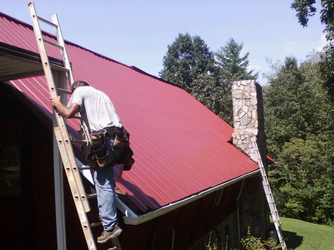 Roofer on a ladder beside a red metal roof and stone chimney — Soffit & Fascia Repair by Roofing Brothers in Franksville
